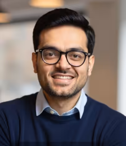 A man with short dark hair, wearing glasses, a blue sweater, and a collared shirt, smiles while facing the camera in an indoor setting.