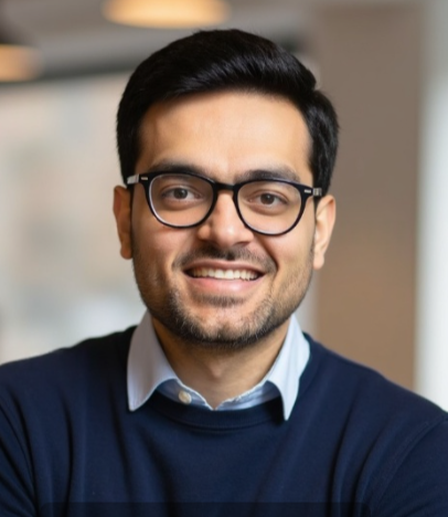 A man with short dark hair, wearing glasses, a blue sweater, and a collared shirt, smiles while facing the camera in an indoor setting.
