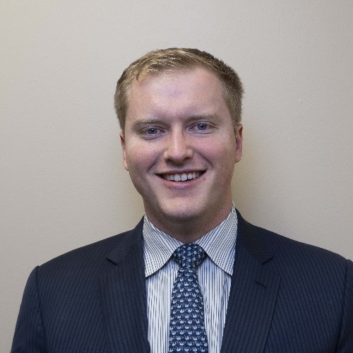 A man in a dark pinstripe suit, light striped shirt, and patterned tie stands in front of a plain beige wall, smiling at the camera.