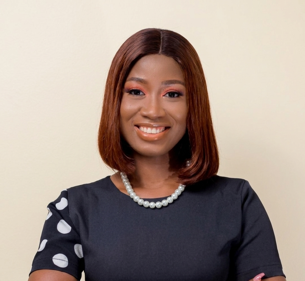 A woman with straight, shoulder-length hair, wearing a black dress with white dots on one sleeve and a pearl necklace, smiles in front of a plain light-colored background.
