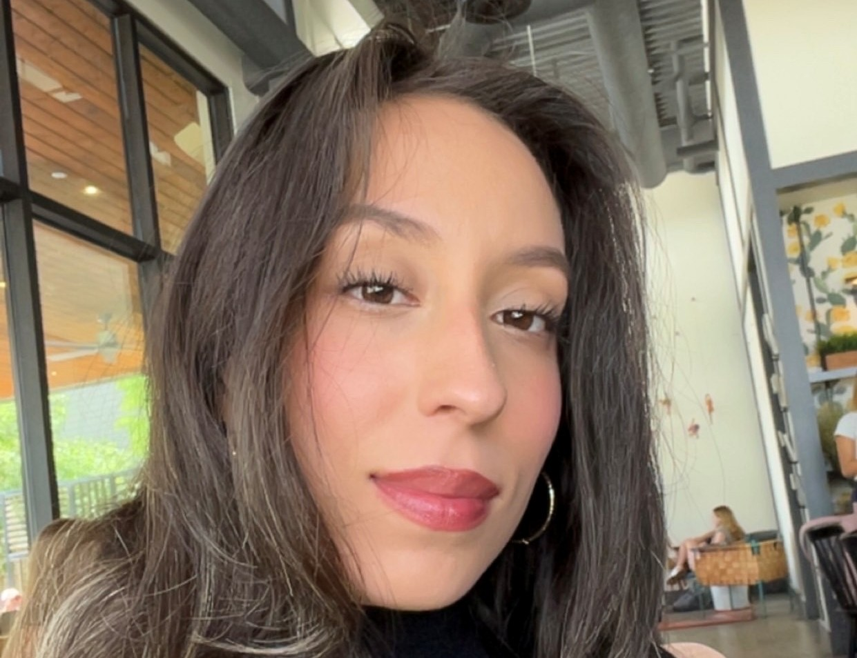 A woman with long dark hair and hoop earrings sits indoors near large windows, looking at the camera.