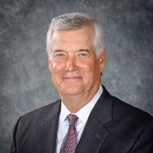 Smiling older man with short gray hair, wearing a dark suit, white shirt, and patterned tie, posed against a gray mottled background.