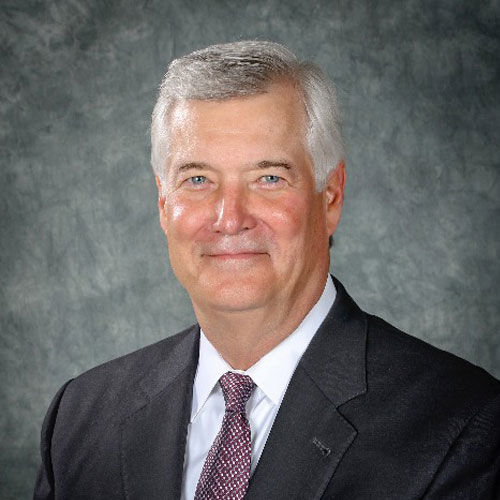 Smiling older man with short gray hair, wearing a dark suit, white shirt, and patterned tie, posed against a gray mottled background.