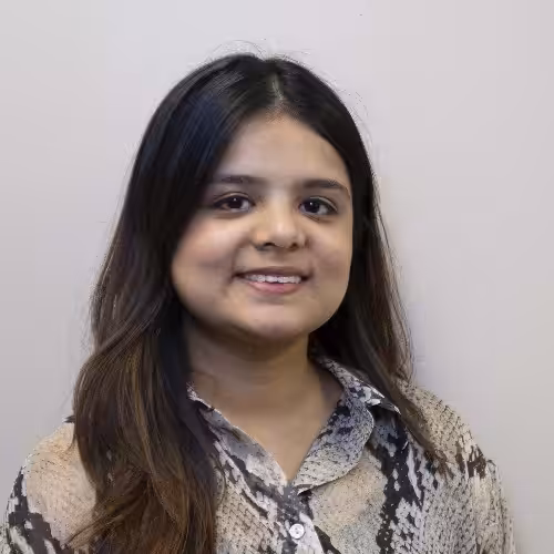 A person with long dark hair and a printed button-up shirt is smiling slightly while facing the camera against a plain light-colored background.