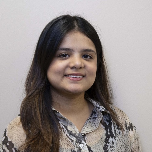 A person with long dark hair and a printed button-up shirt is smiling slightly while facing the camera against a plain light-colored background.