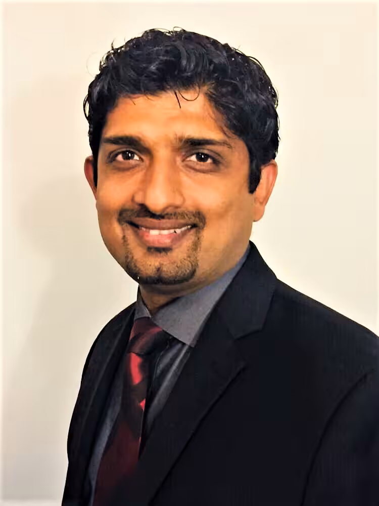 A man in a dark suit, dress shirt, and striped tie is smiling at the camera against a plain light background.
