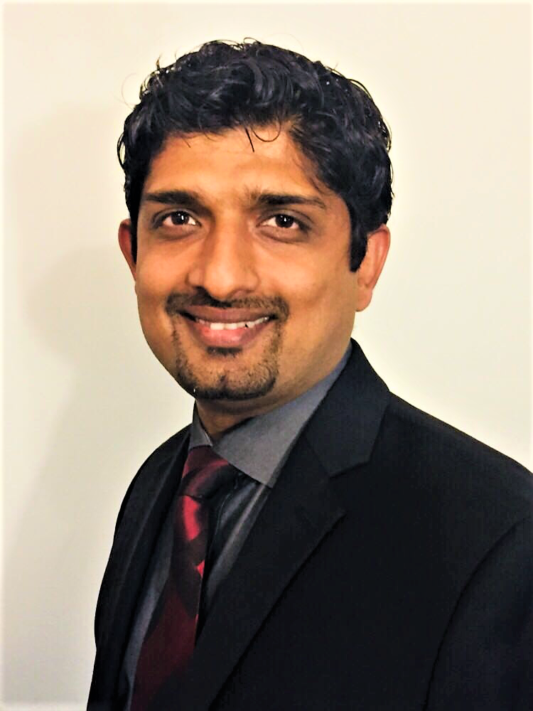 A man in a dark suit, dress shirt, and striped tie is smiling at the camera against a plain light background.
