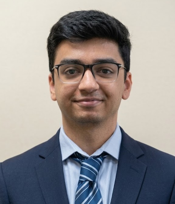 A man wearing glasses, a dark suit, a light blue shirt, and a blue striped tie stands in front of a plain beige background.